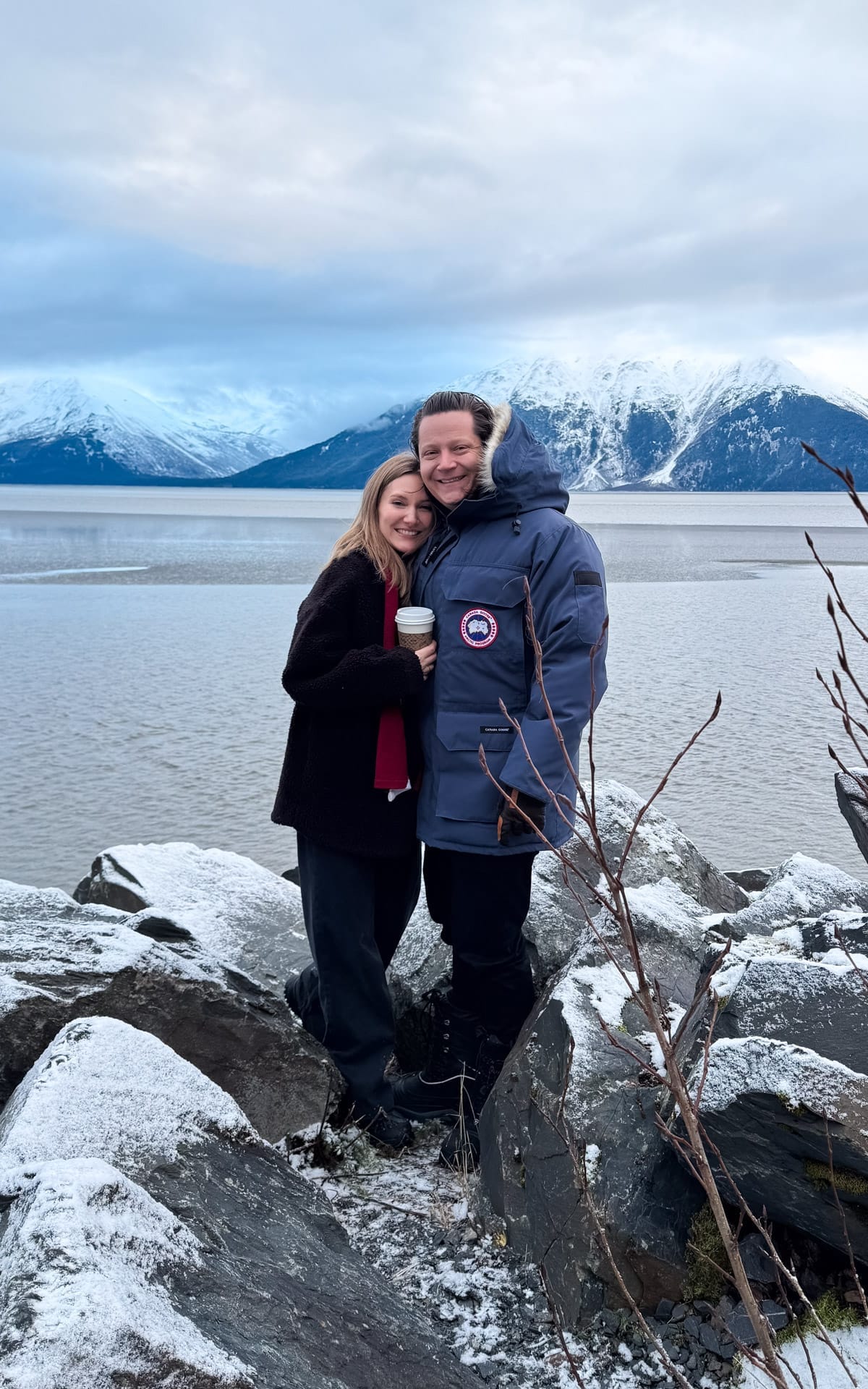 Cec and Ari standing on snowy shoreline rocks along Turnagain Arm with snow-covered mountains and calm water in the background.