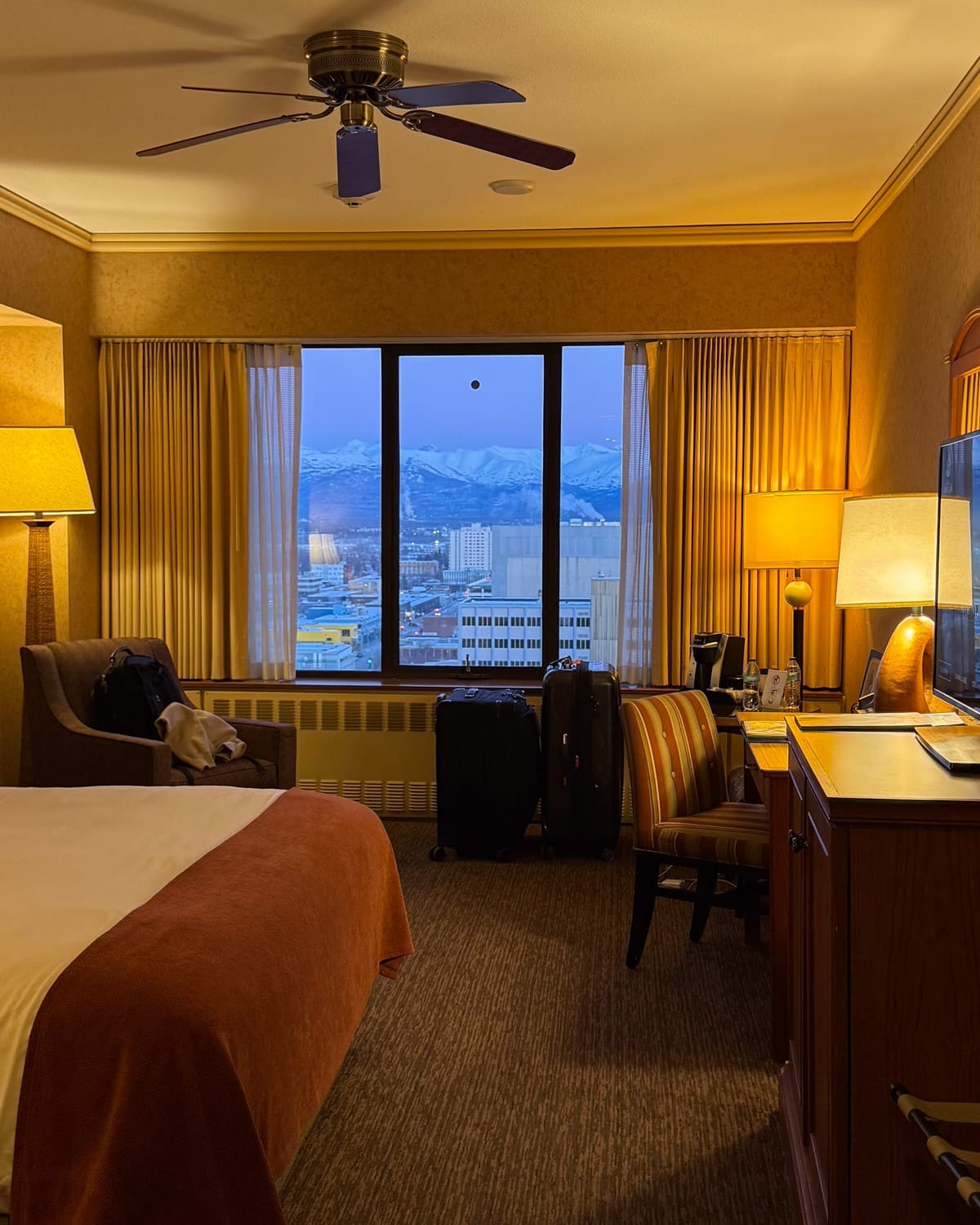 Room with warm lighting at Hotel Captain Hook in downtown Anchorage with large window overlooking snow-covered mountains.