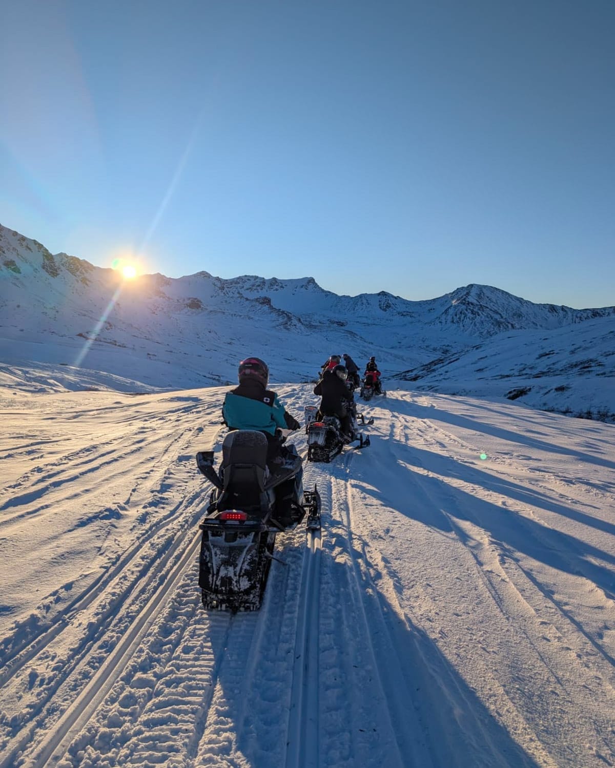 Group of snowmobilers riding through a wide alpine valley near Hatcher Pass at sunset, with snowy peaks and long winter shadows.