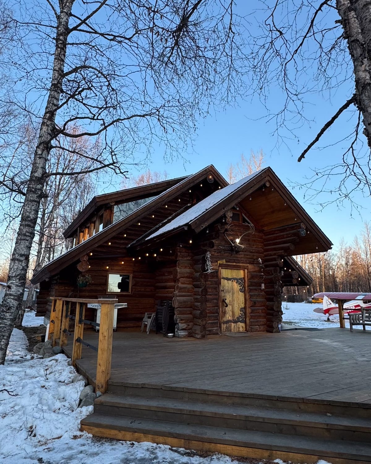 Exterior of a two-story log cabin in Talkeetna with wooden deck and small bush plane parked nearby.