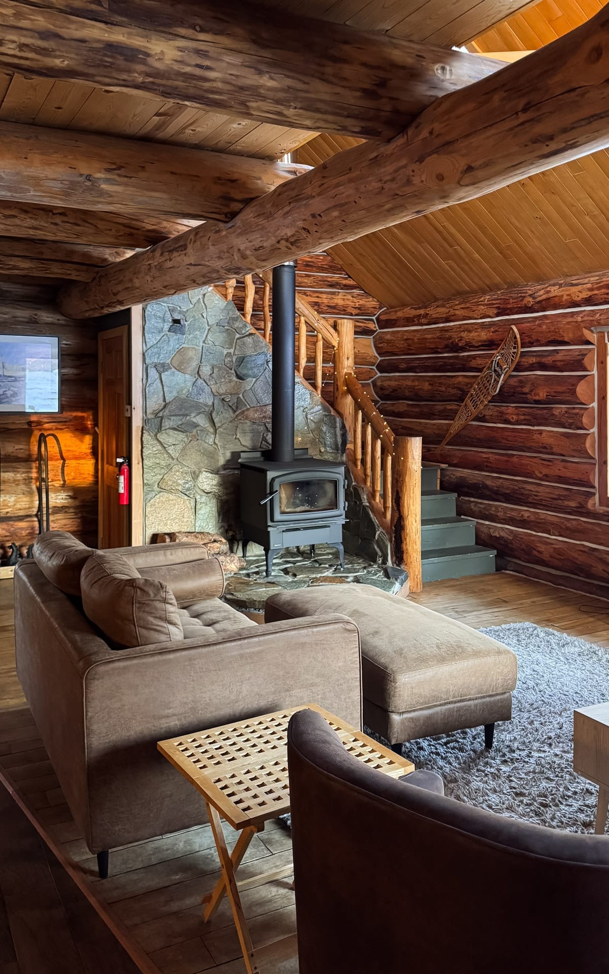 Cozy log cabin living room with wood-burning stove, stone fireplace surround, and staircase leading to loft.