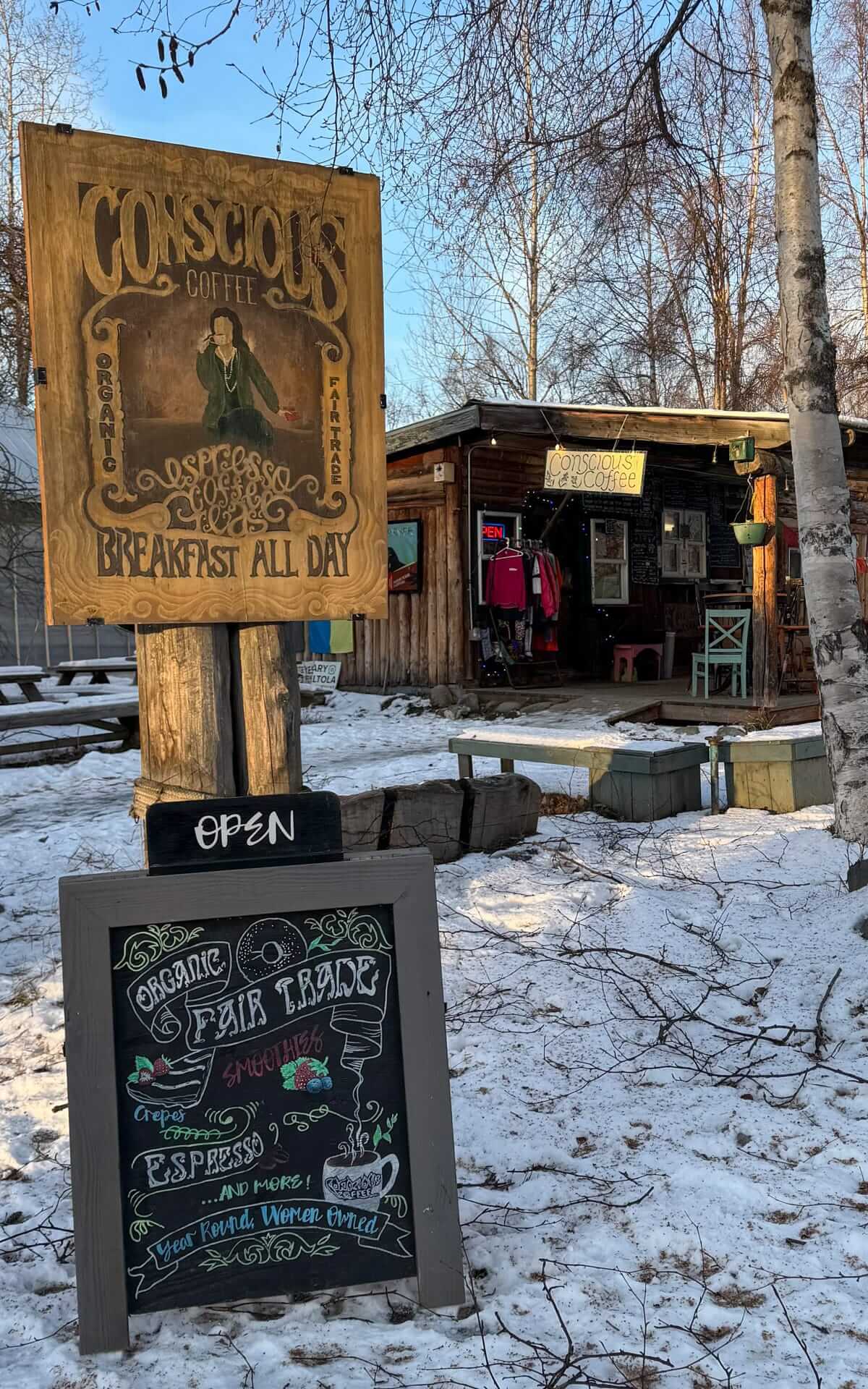 Rustic wooden exterior of Conscious Coffee in Anchorage, surrounded by snow and birch trees, with hand-painted espresso and breakfast signage.