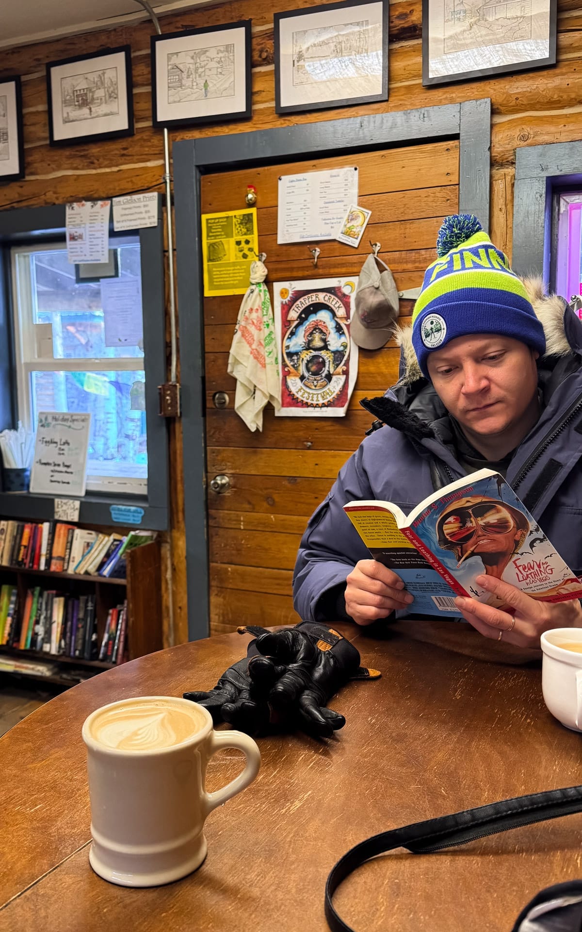 Latte on wooden table beside Ari reading Fear and Loathing in Las Vegas at Conscious Cafe in Talkeetna.