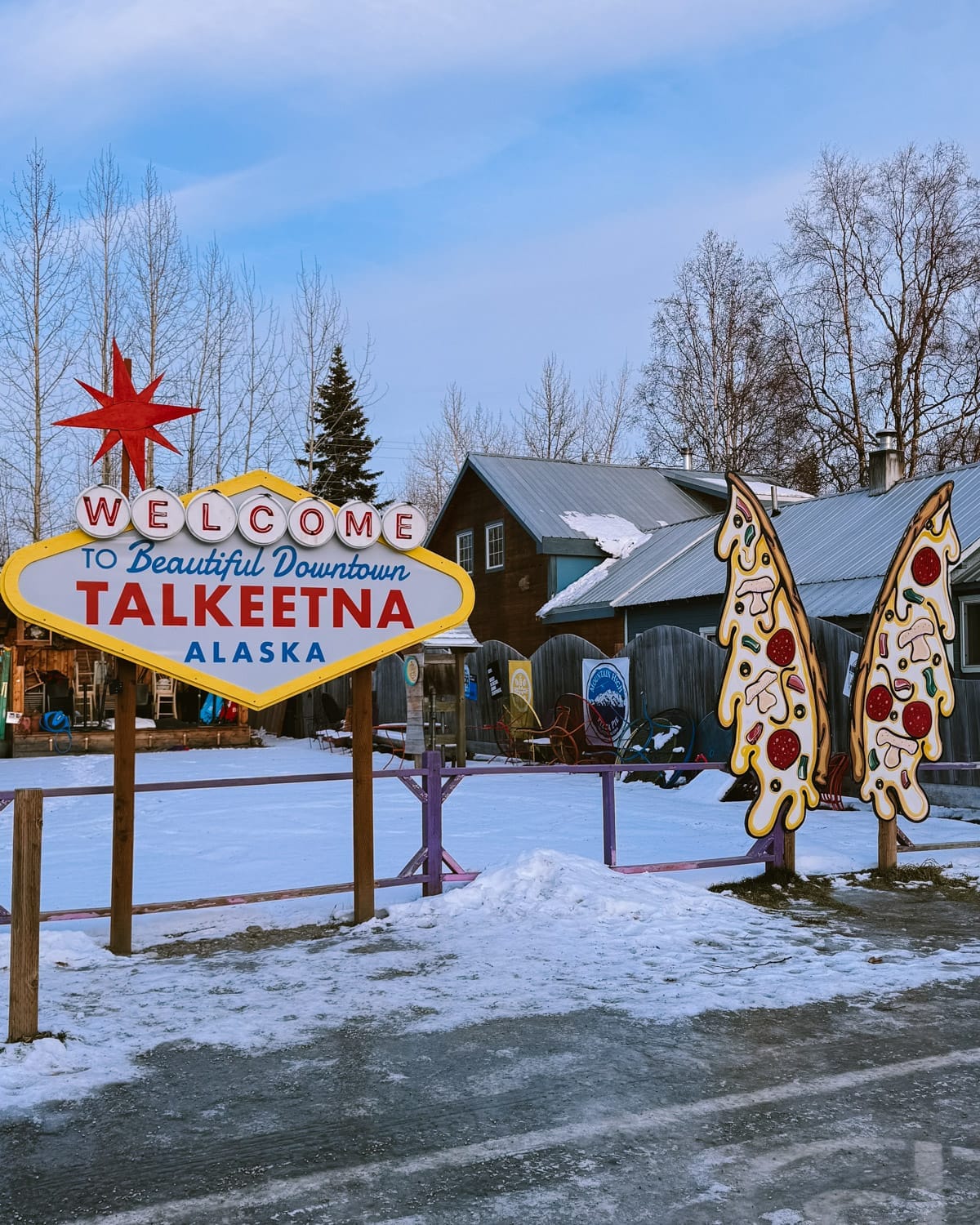 A Vegas style Welcome to Talkeetna sign with snow on the ground and quirky pizza sculptures beside it.