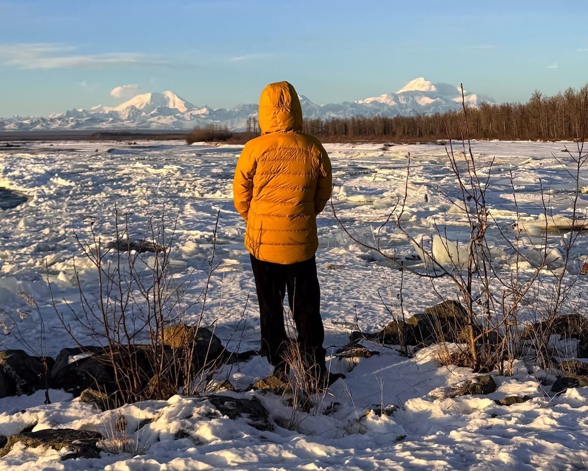 Cec in yellow jacket standing on a snowy riverbank looking toward Denali and surrounding mountain peaks at golden hour.