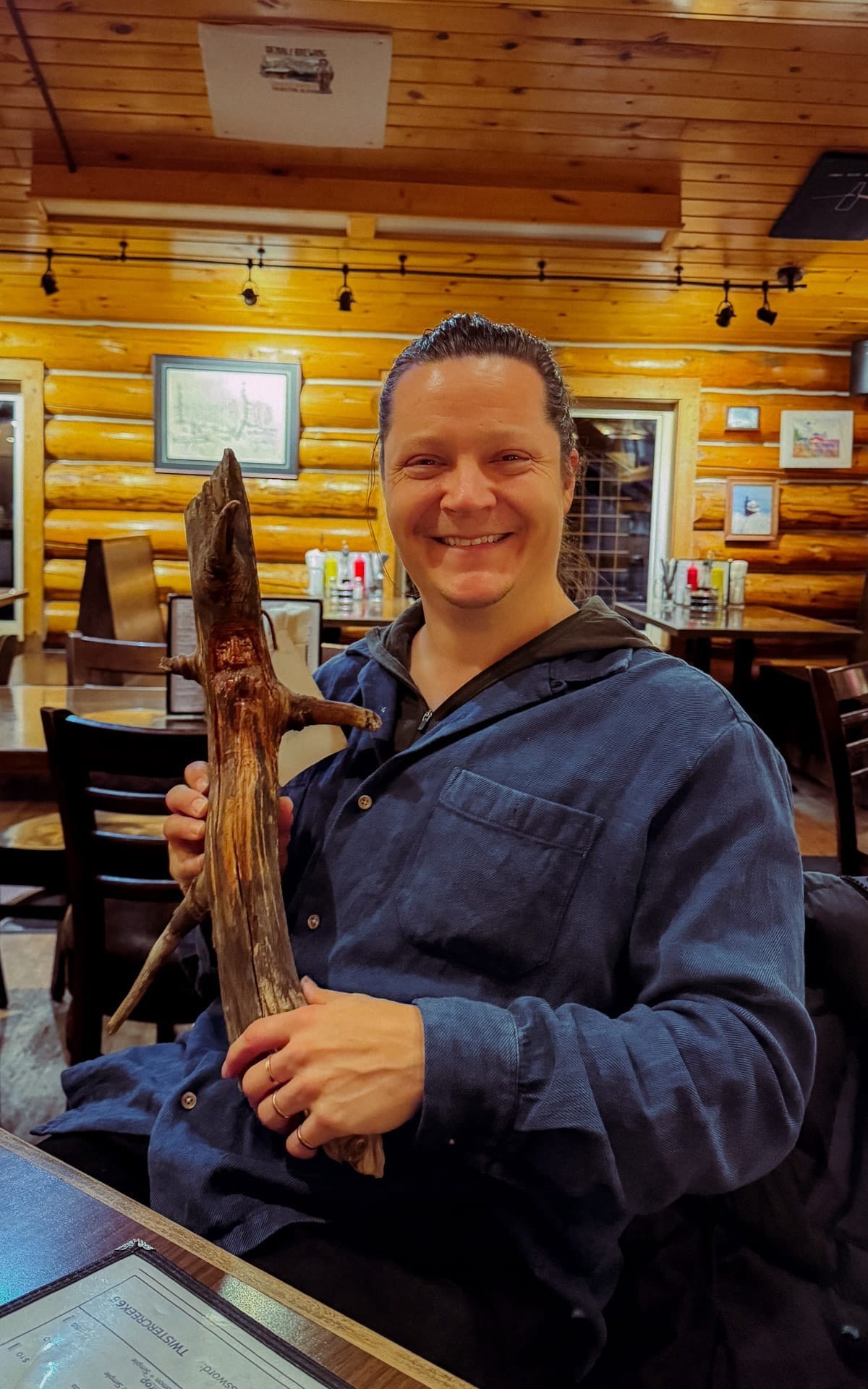 Ari smiling holding a hand-carved wooden branch sculpture inside a cozy log cabin restaurant with timber walls and warm lighting in Talkeetna.