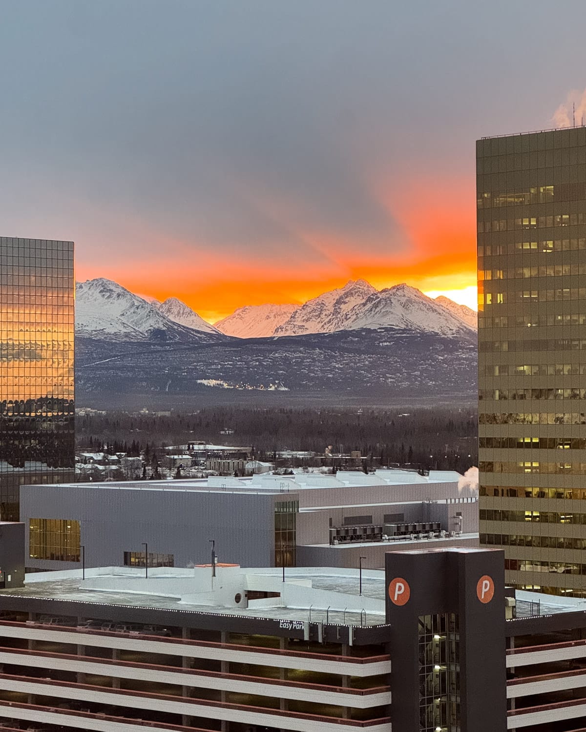 Sunset over downtown Anchorage with snow-covered Chugach Mountains glowing orange between glass office towers.