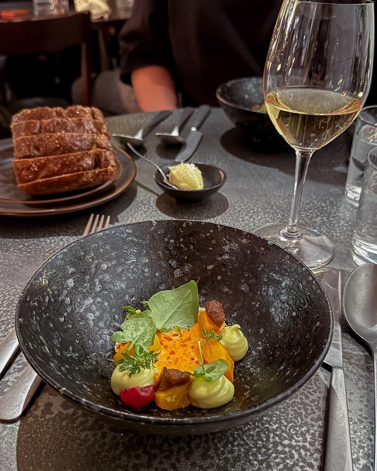 Modern Nordic fine-dining dish served in a black bowl, with sourdough bread and white wine on a table at Plein in Helsinki..