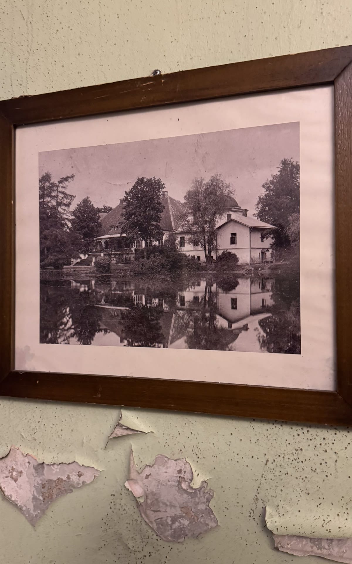Framed vintage photograph of the Maidla manor house in its golden years, reflected in a pond, hanging on cracked plaster wall.