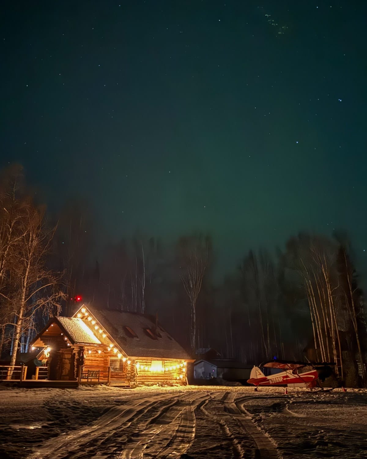 Log cabin in Talkeetna lit up at night with a small red bush plane parked beside it and the northern lights glowing overhead.