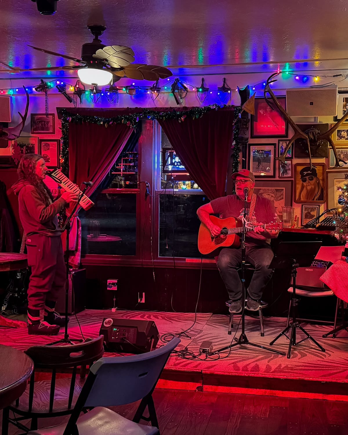 Two musicians performing live music on a small stage in a colourful, neon-lit Talkeetna Inn decorated with framed photos and antlers.