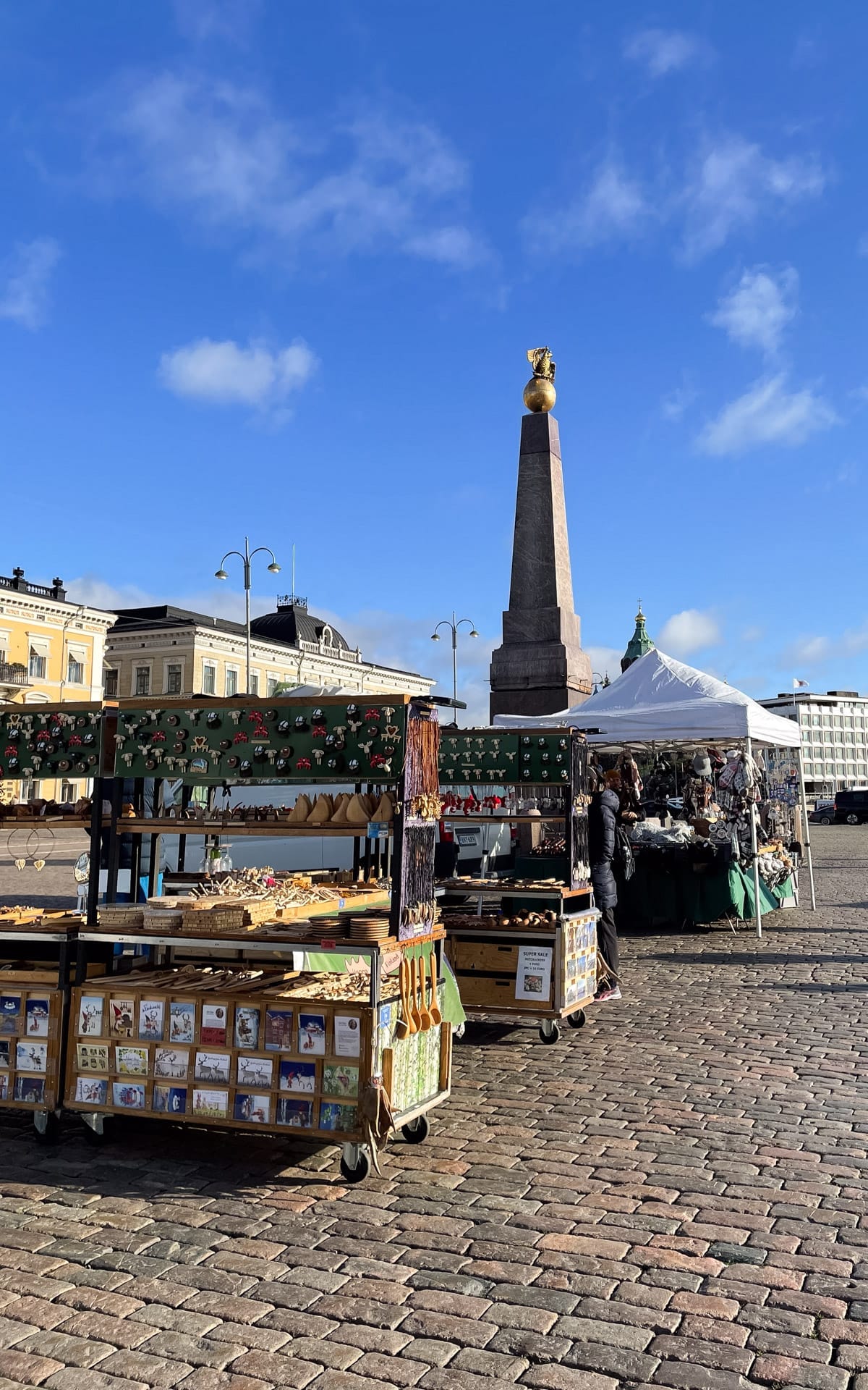 Market stalls at Helsinki’s Market Square with handmade souvenirs and the obelisk monument rising behind.