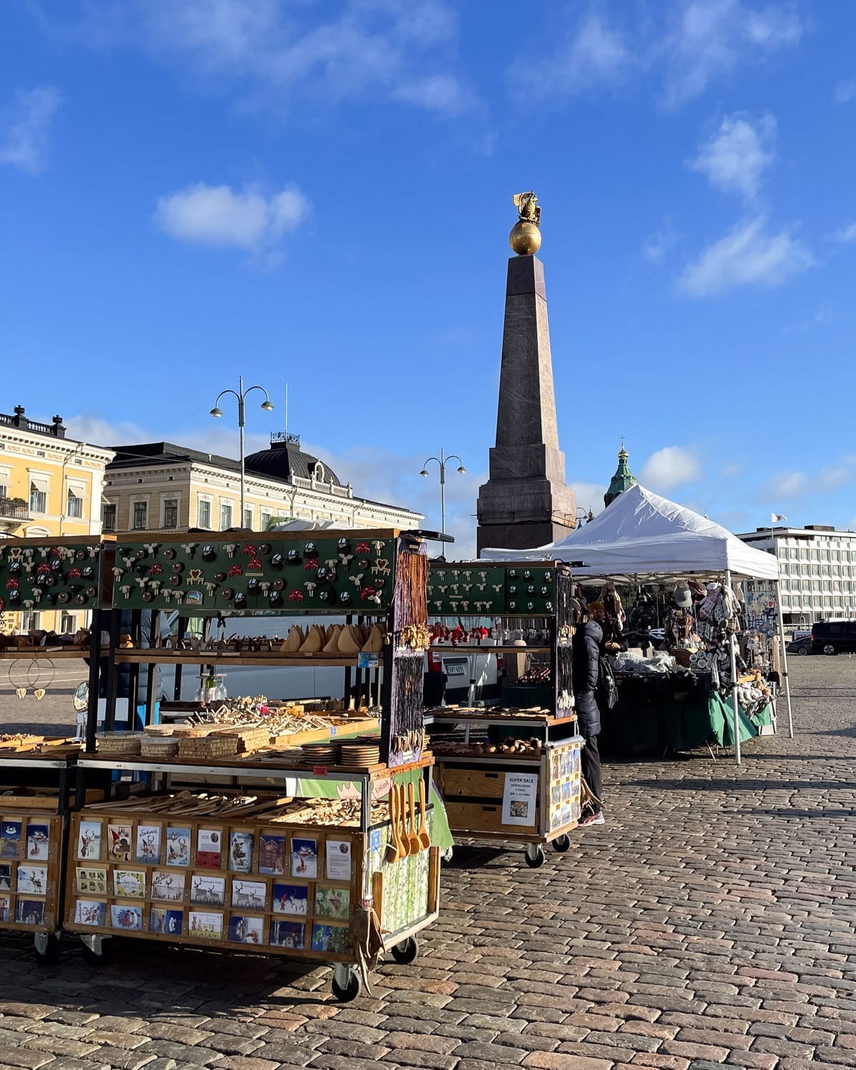 Market stalls at Helsinki’s Market Square with handmade souvenirs and the obelisk monument rising behind.