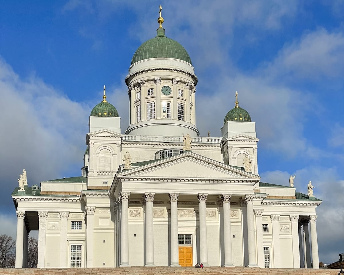 Helsinki Cathedral with its white neoclassical façade and green domes rising above the steps under a blue sky.