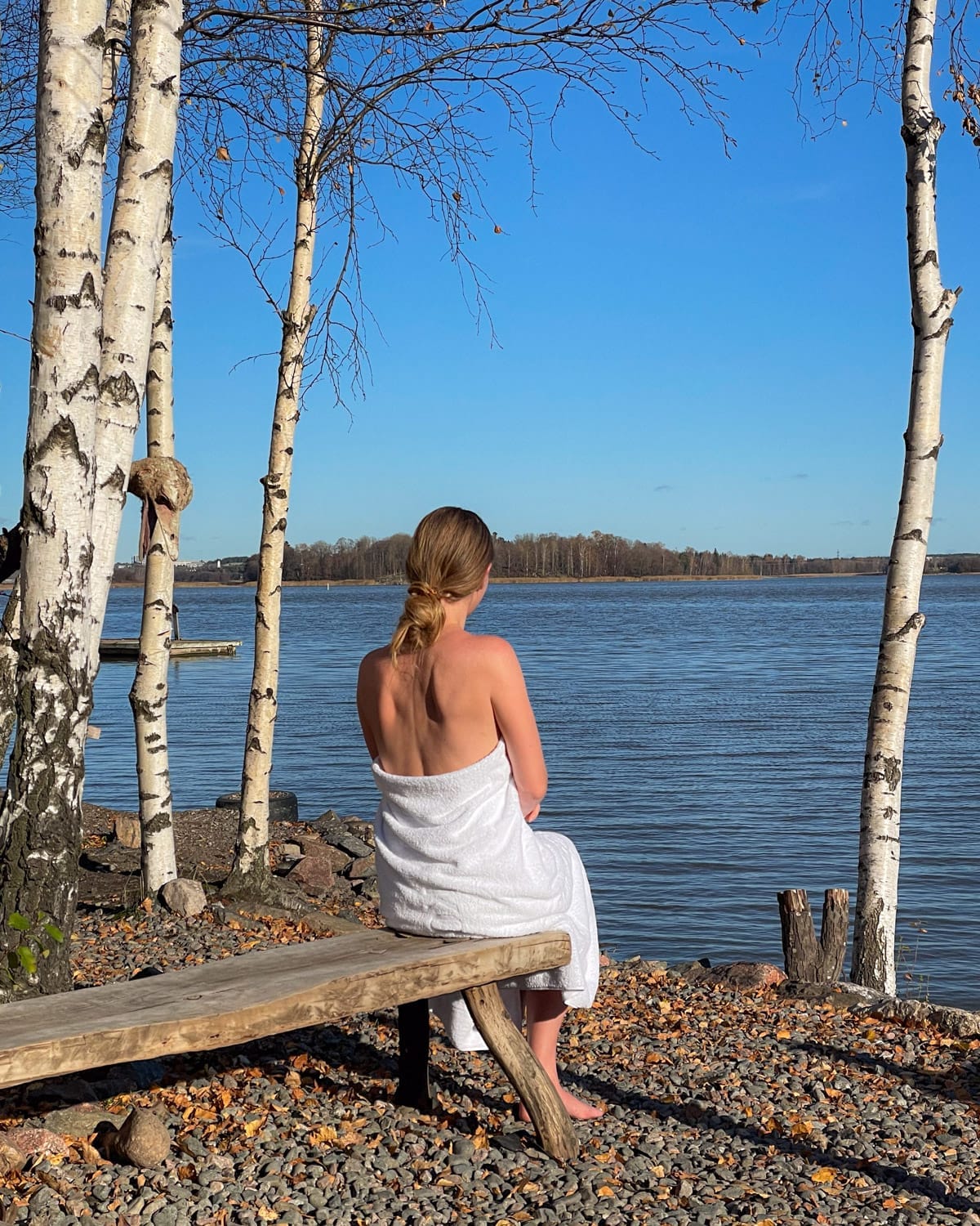 Cec wrapped in a towel sitting by the sea between birch trees at Sompasauna.