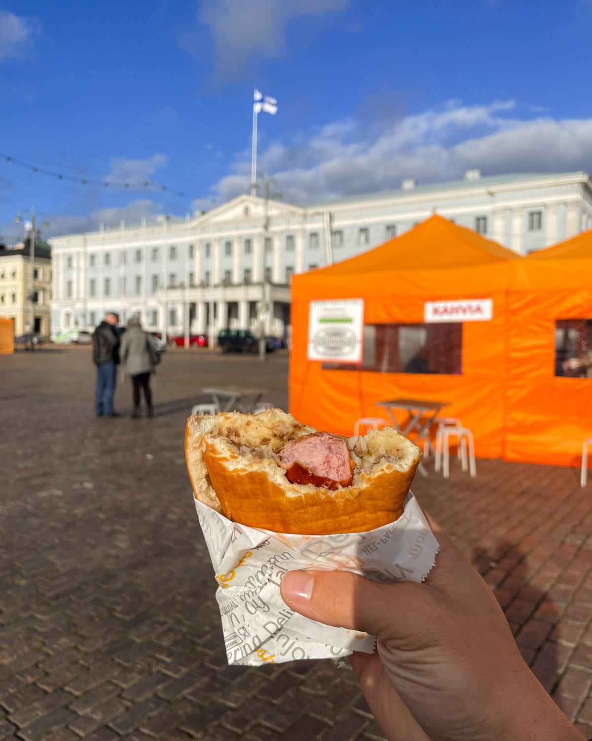 Lihapiirakka meat pie with a hot dog and mustard held in front of the bright orange coffee tent at Helsinki Market Square.