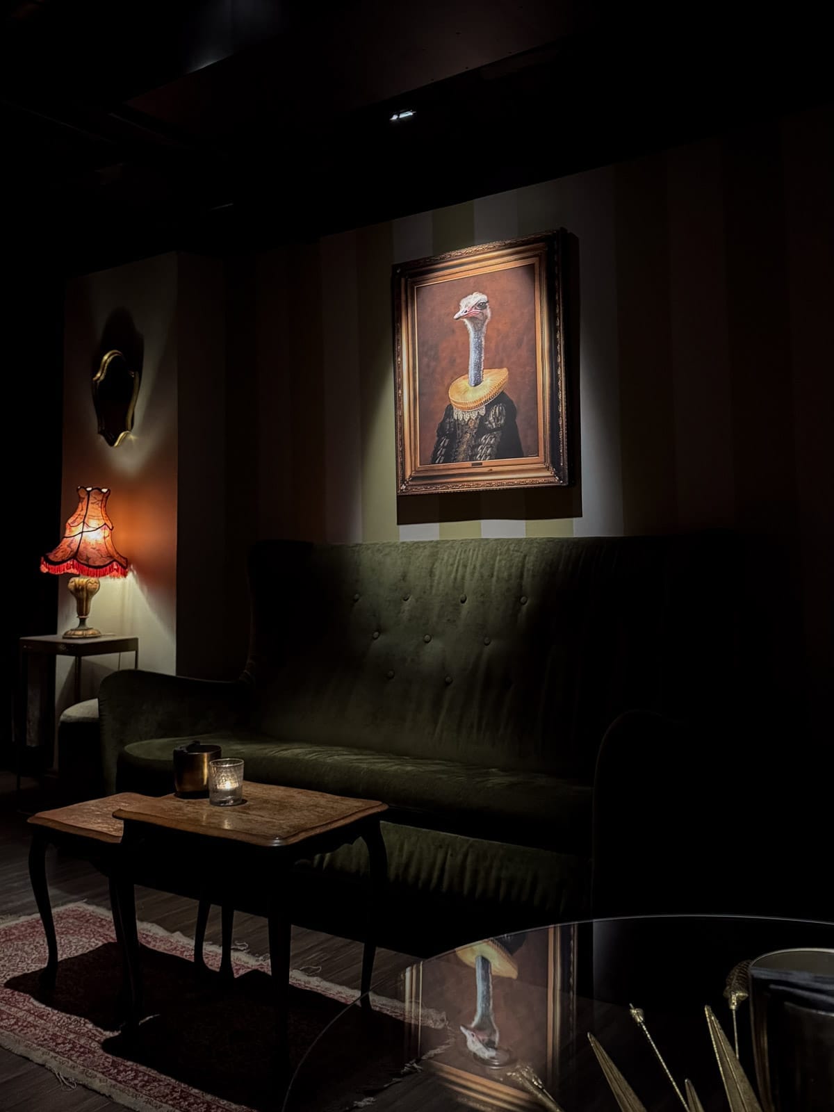 Moody cocktail lounge with velvet green sofa, vintage lamp and framed ostrich portrait at Whisper Sister.