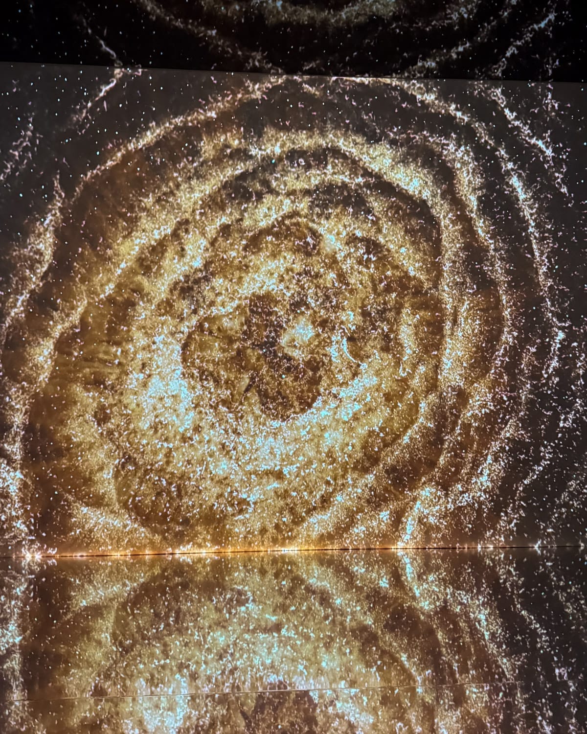 Immersive golden galaxy light installation reflected on a mirrored floor in a dark exhibition space at Fotografiska Tallinn.