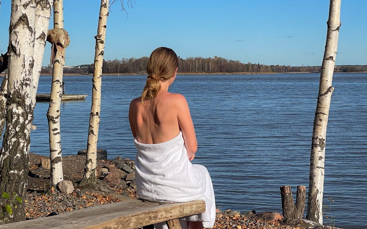 Cec wrapped in a towel sitting by the sea between birch trees at Sompasauna, a stop in the 2 day Helsinki itinerary.