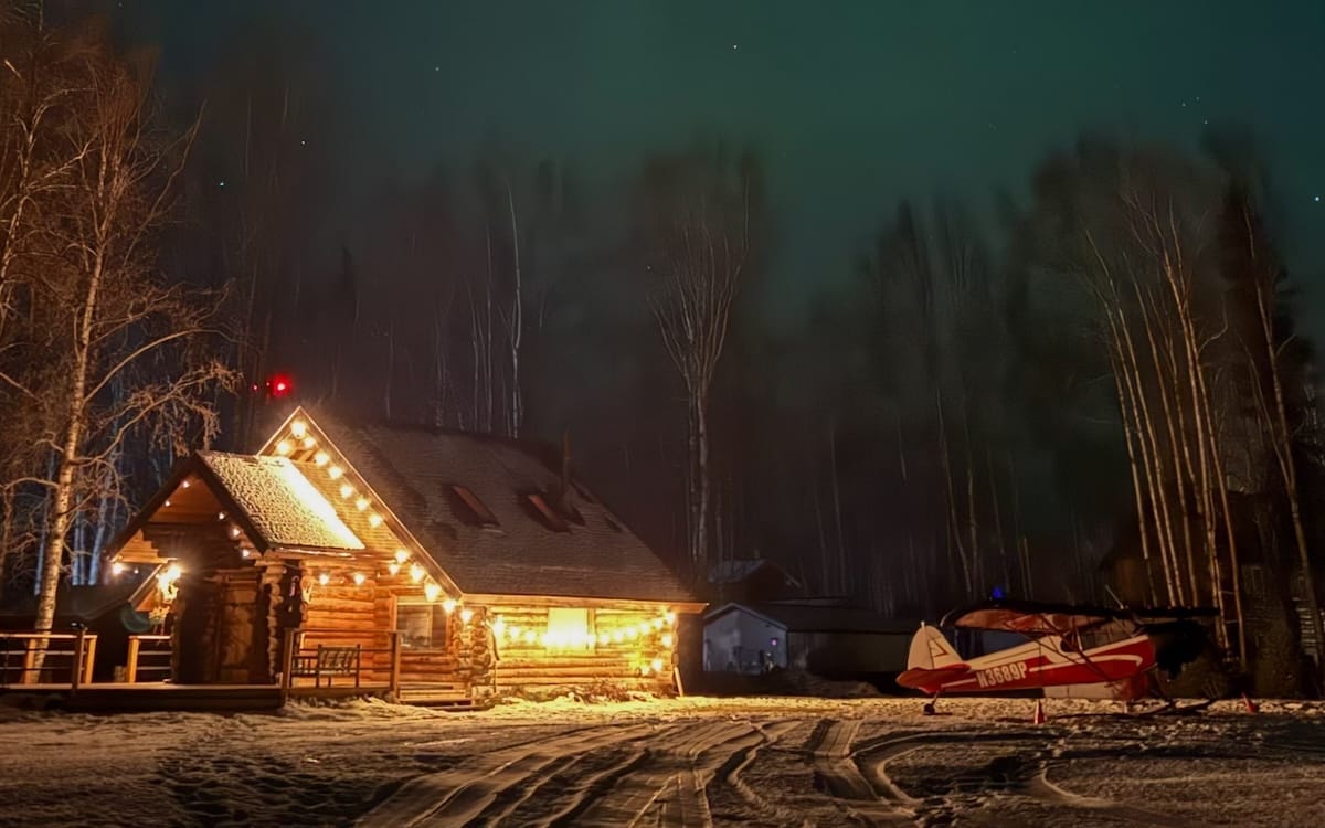 Log cabin lit up at night with the northern lights overhead in Talkeetna, a stop on the 5 Day Alaska Itinerary.